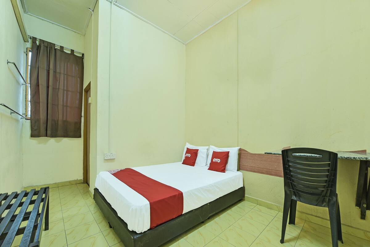 A single bed with a white and red bedspread, two pillows with 'OYO' branding, a wooden headboard, a small table, a chair, and a window with curtains.