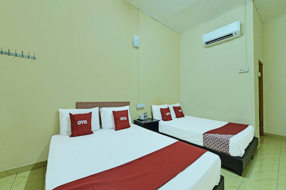 A hotel room with two beds, each with red and white bedding and pillows, a nightstand, an air conditioning unit, and a tiled floor.