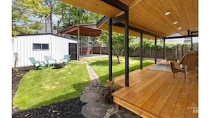 Ein überdachter Patio-Bereich mit Holzterrasse, Sitzgelegenheiten im Freien und einem kleinen, weißen Gebäude im Hintergrund.