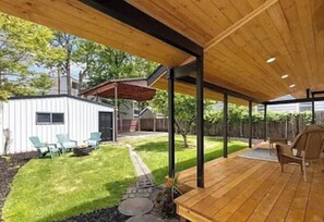Un patio couvert avec une terrasse en bois, des sièges d’extérieur et un petit bâtiment blanc en arrière-plan.