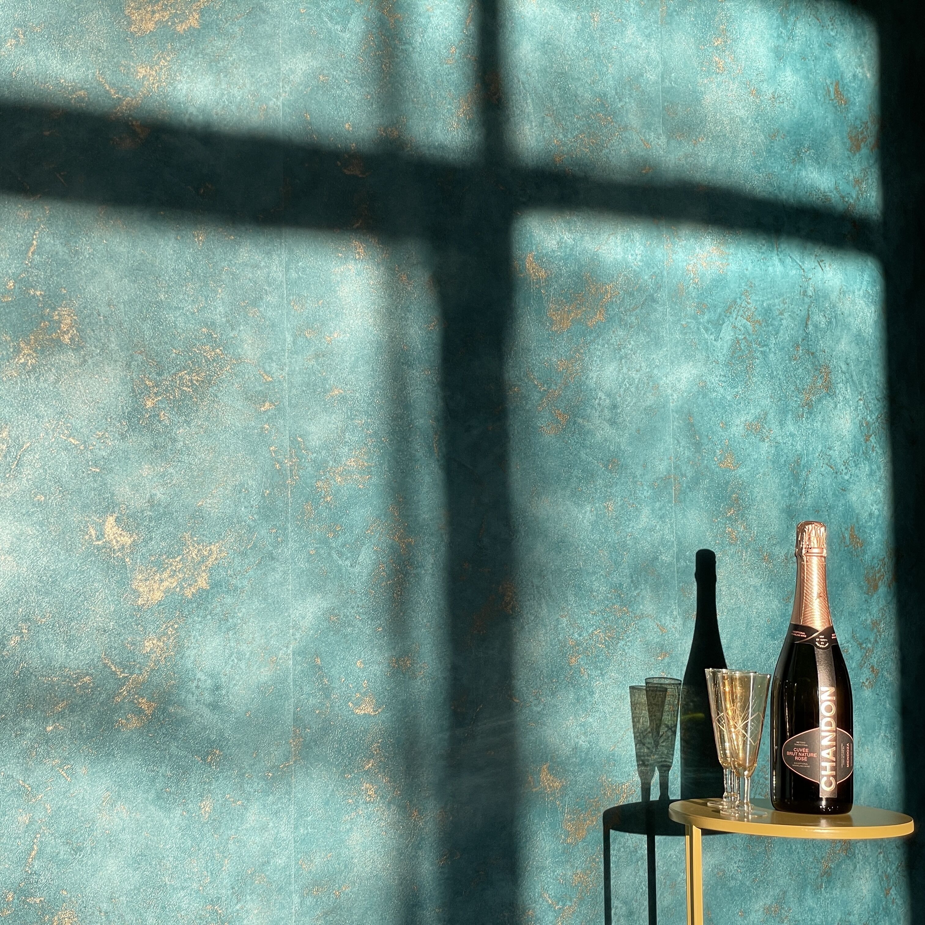 A table with a bottle of champagne and a glass, set against a textured teal wall with a shadow cast across it.