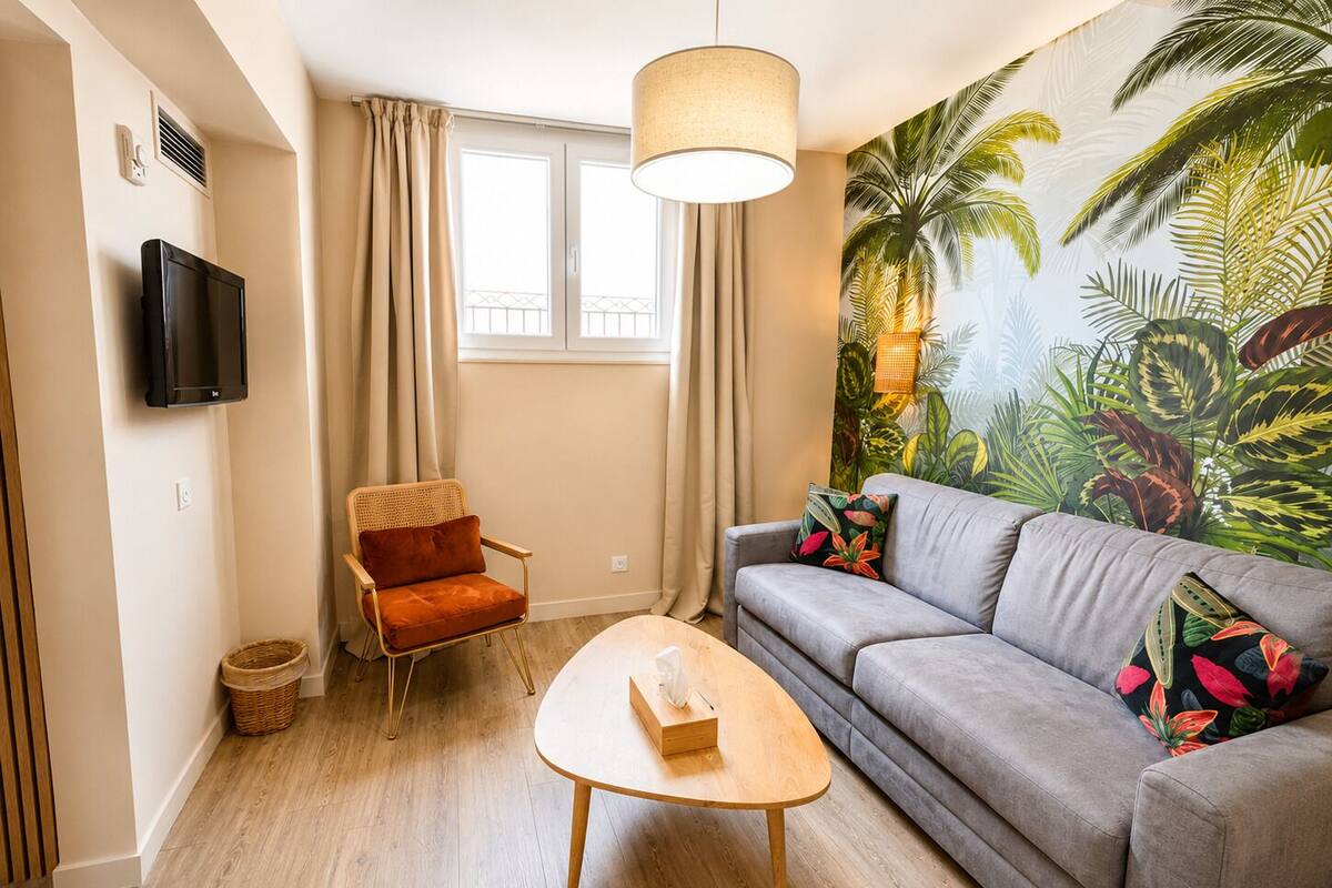 A living room with a sofa, a chair, a coffee table, a TV, and a large tropical mural.