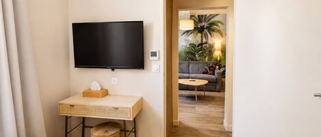 Habitación de hotel con cama, televisor montado en la pared, una pequeña mesa de madera con una caja de pañuelos y una puerta que da a una sala de estar con sofá y lámpara.