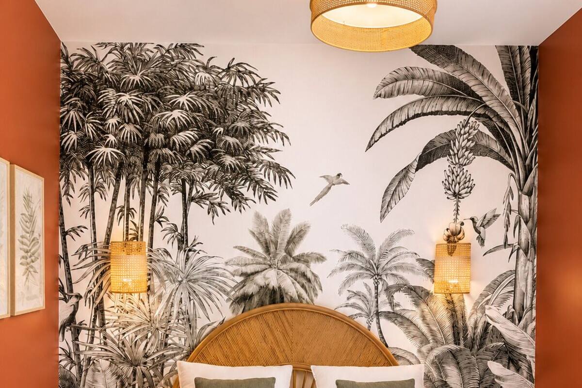 A bedroom with a bed, a wall mural of tropical plants, two wall-mounted lamps, and a round wooden stool.