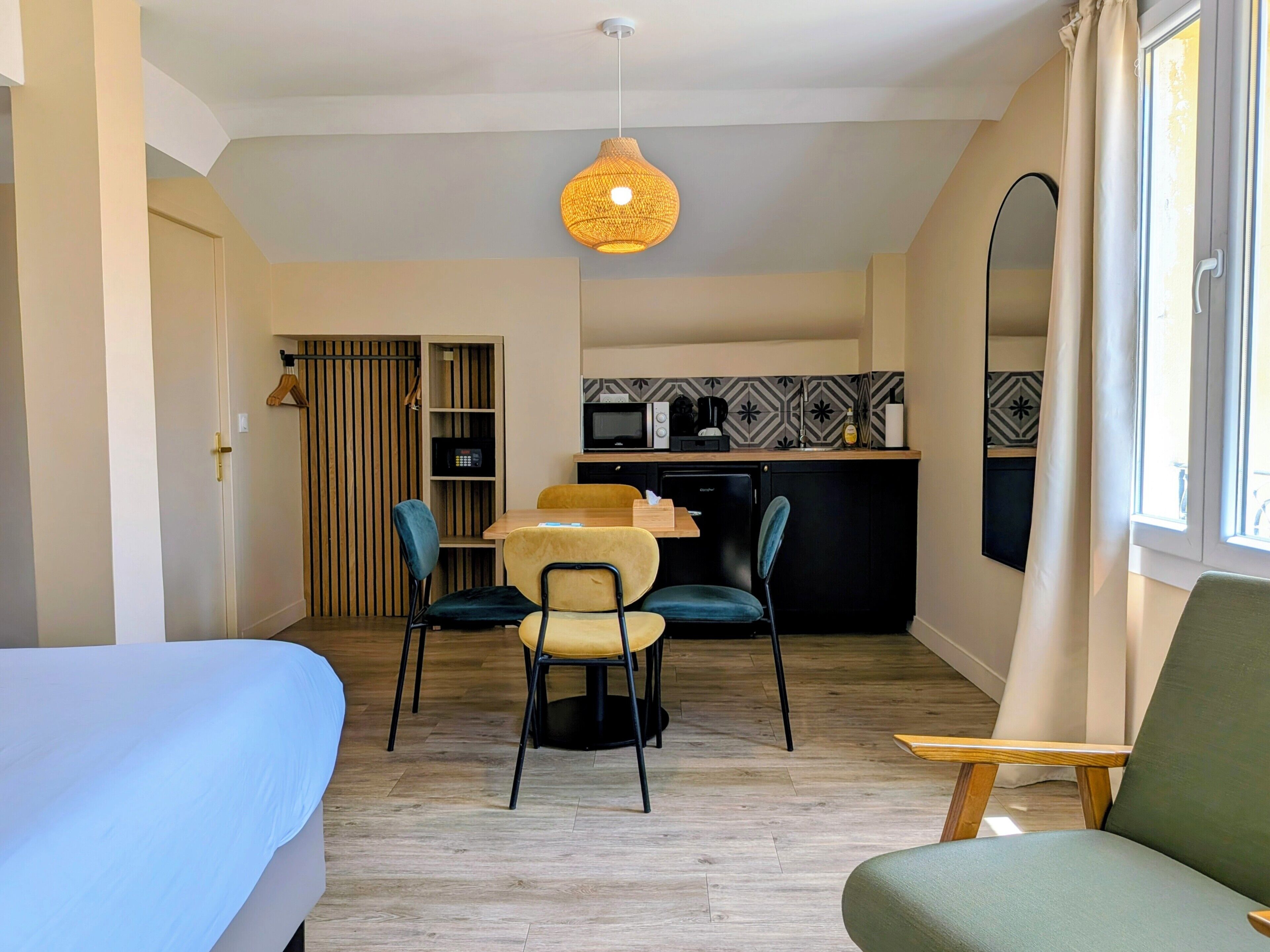 A compact room with a bed, a dining table with chairs, a kitchenette, and a large mirror.
