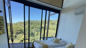 A modern bedroom with two beds, a bench, and a large sliding glass door leading to a balcony with a forest view.