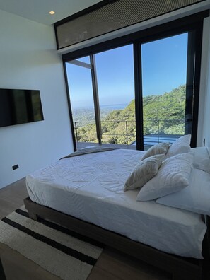 A modern bedroom with a large bed, a view of a mountainous landscape, and a flat-screen TV mounted on the wall.
