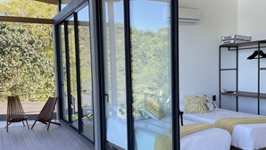 A modern bedroom with a large sliding glass door leading to a balcony, two beds with white linens, a wooden ceiling, and a view of a forested area.