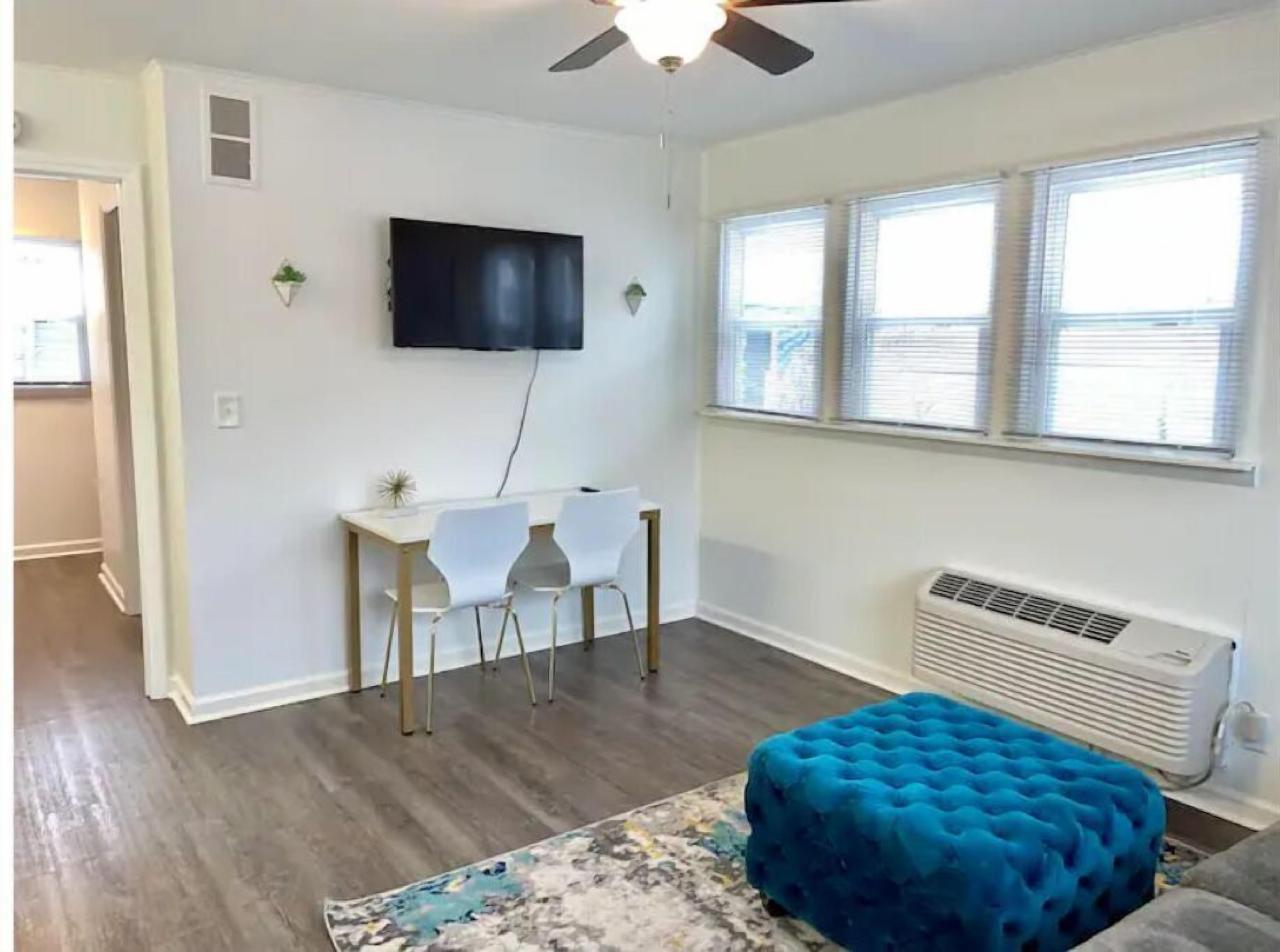 A living room with a flat-screen TV mounted on the wall, a small table with two white chairs, a blue ottoman, and a window with blinds.