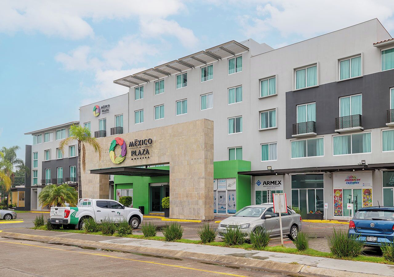 Un hotel de varios pisos con un diseño moderno, que cuenta con un llamativo letrero de 'MAEICO PLAZA', una farmacia y un concesionario de automóviles.