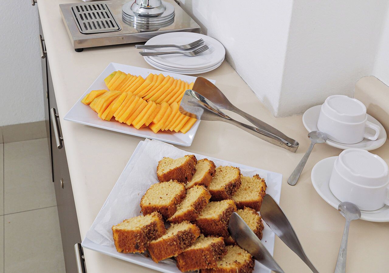 Um buffet de café da manhã com fruta fatiada e bolo, talheres e copos brancos sobre uma mesa.