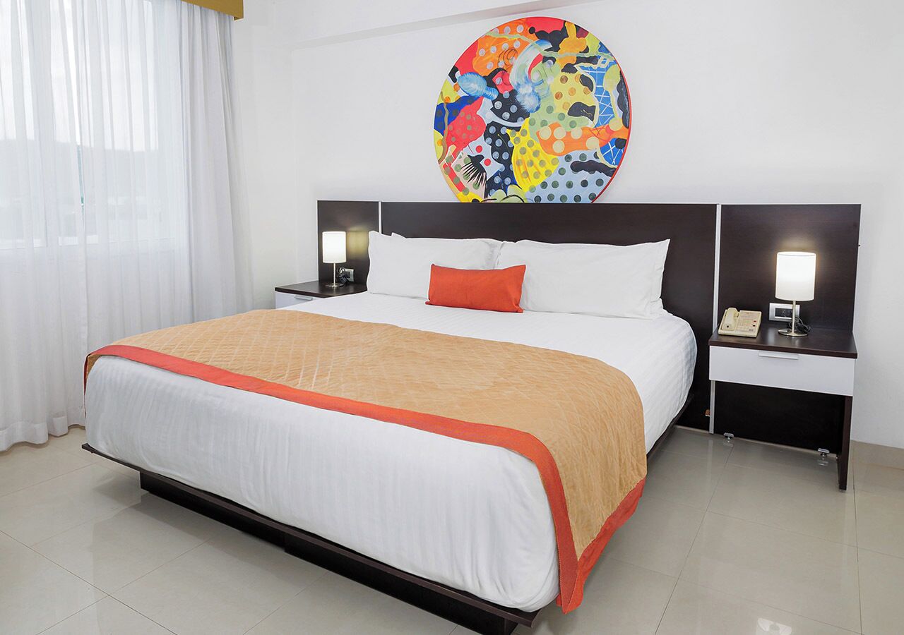 A hotel room with a large bed, white bedding, an orange throw, a colorful abstract painting on the wall, and two bedside tables with lamps and a phone.