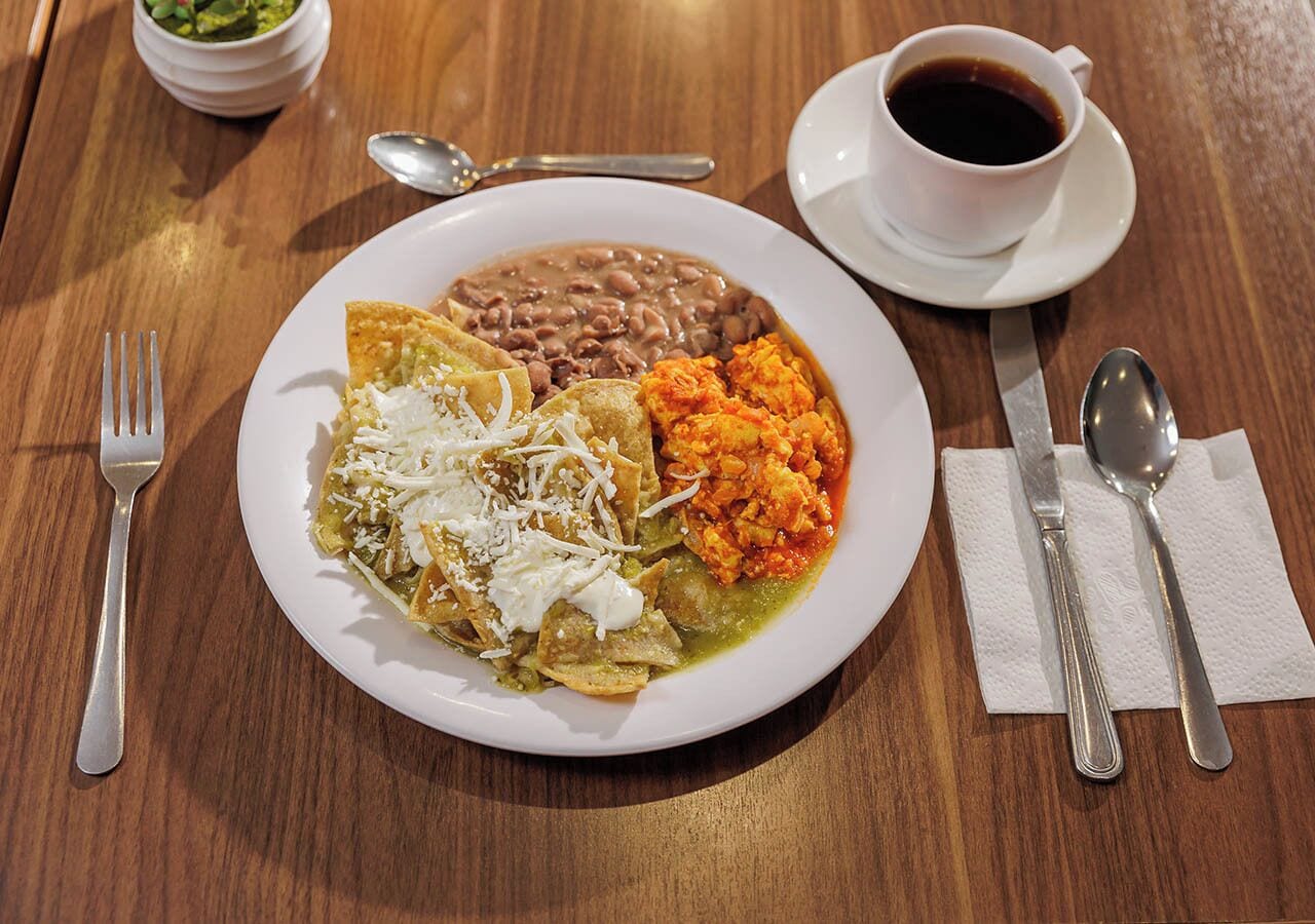 A plate of food with beans, refried beans, and a green sauce, accompanied by a cup of coffee and cutlery on a wooden table.