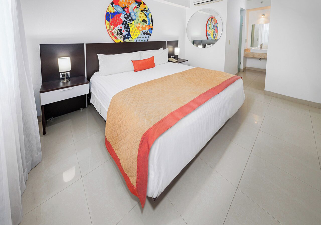 A hotel room with a large bed, a nightstand with a lamp, a round mirror, and a colorful artwork on the wall.