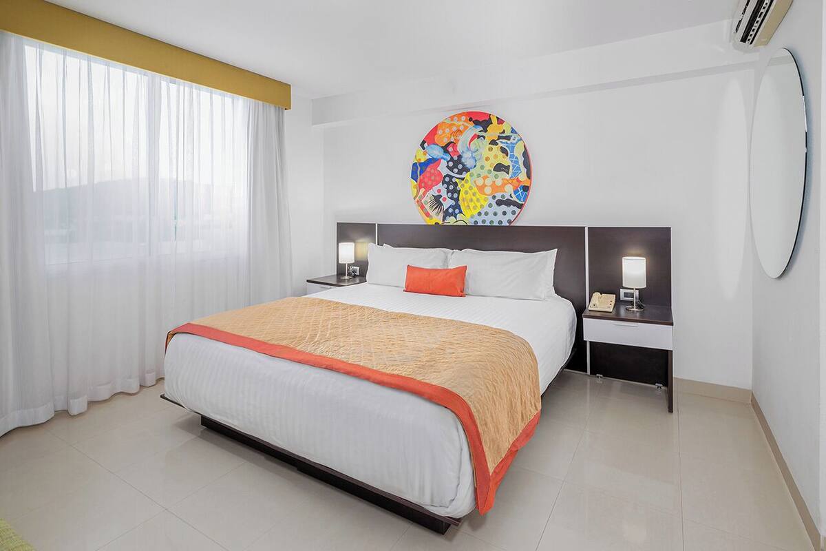 Habitación de hotel moderna con una cama grande, dos mesitas de noche, una obra de arte circular en la pared y un ventanal con cortinas translúcidas.