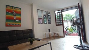 A room with a black leather sofa, a wooden coffee table, a small table with a speaker, and a wall-mounted fan. There is a window with a view of greenery outside. - Beautiful small house close Getsemani (Cartagena de Indias)