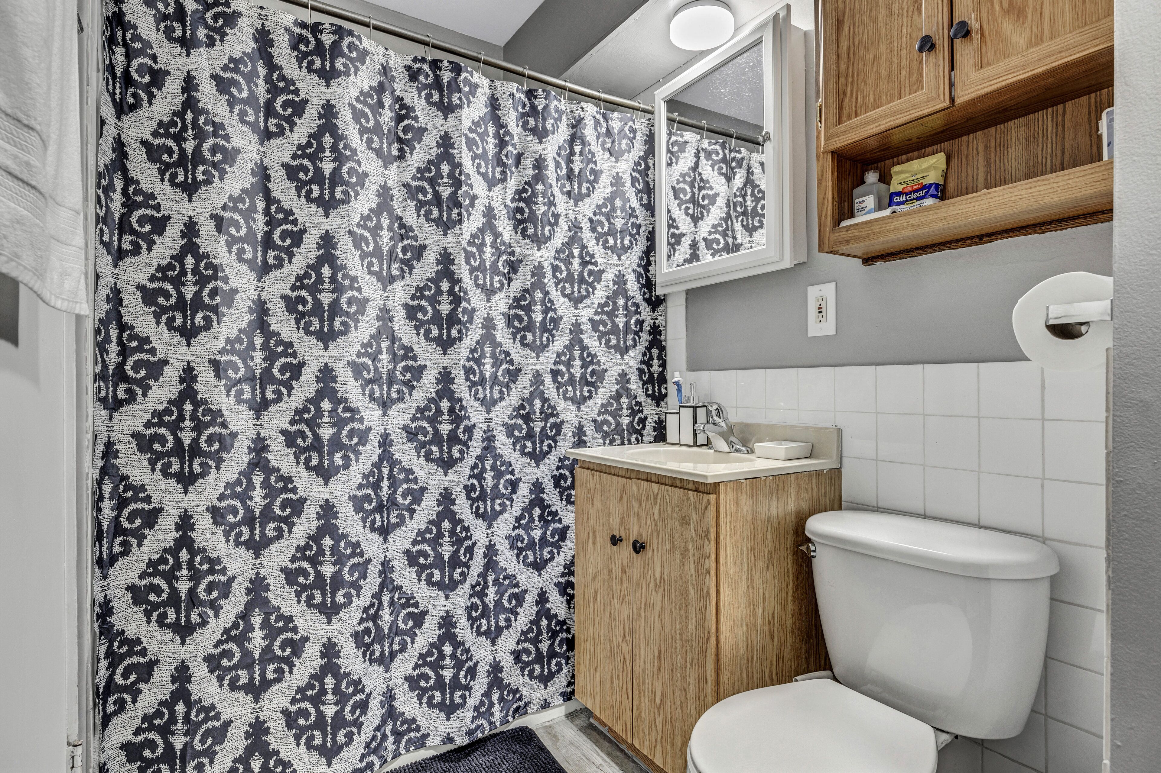 Une salle de bain avec un rideau de douche à motifs, des meubles en bois, des toilettes blanches et un miroir au-dessus du lavabo.