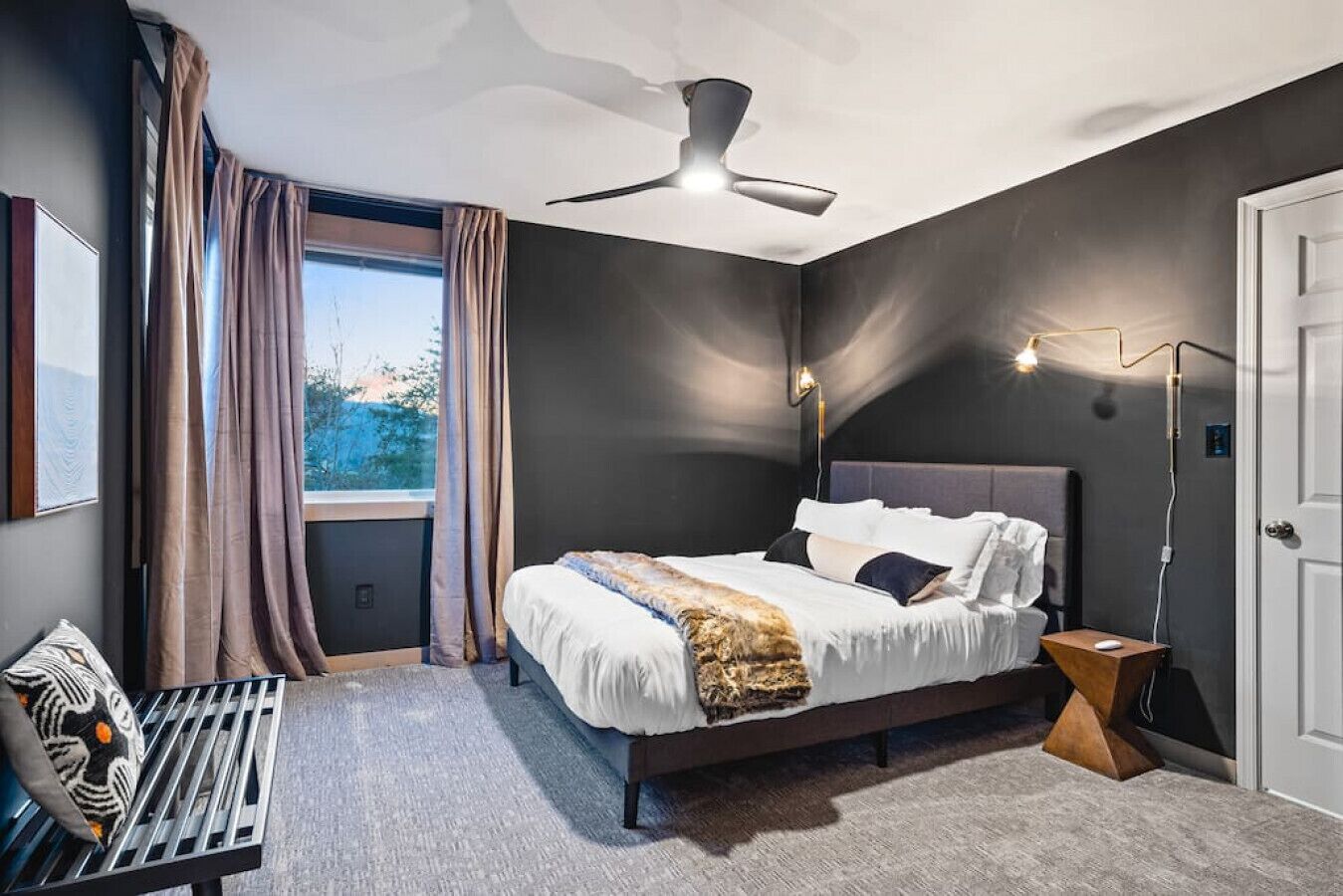 Een moderne slaapkamer met een bed, een plafondventilator, een nachtkastje met een lamp en uitzicht op de omgeving door een raam.