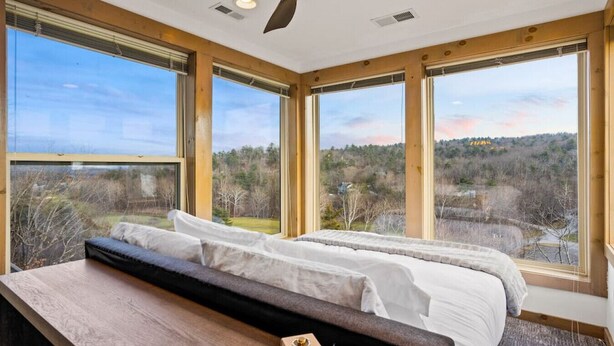 Een slaapkamer met een groot bed, een houten bureau en uitzicht op een bebost landschap door grote ramen.