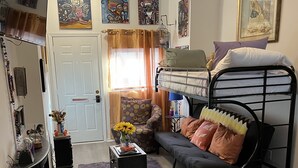 A cozy living room with a bunk bed, a sofa, a chair, and a small table. There are paintings on the walls and a ceiling fan.
