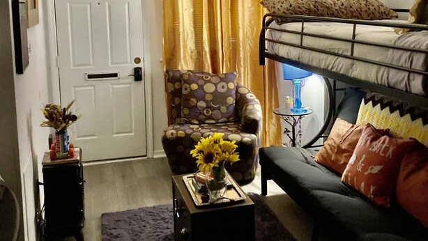 A compact living space with a bunk bed, a sofa, a chair, a coffee table with a vase of flowers, and a door with a mailbox.