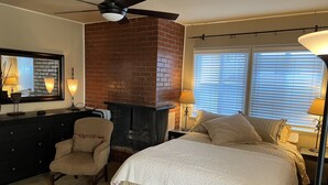 A bedroom with a brick fireplace, a bed with a white quilt, a chair, and a desk with a lamp. - Charming Studio w/ Fireplace & Kitchen — Steps to Sand (Manhattan Beach)