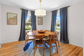 A dining room with a round wooden table, six chairs, a chandelier, and two windows with curtains.