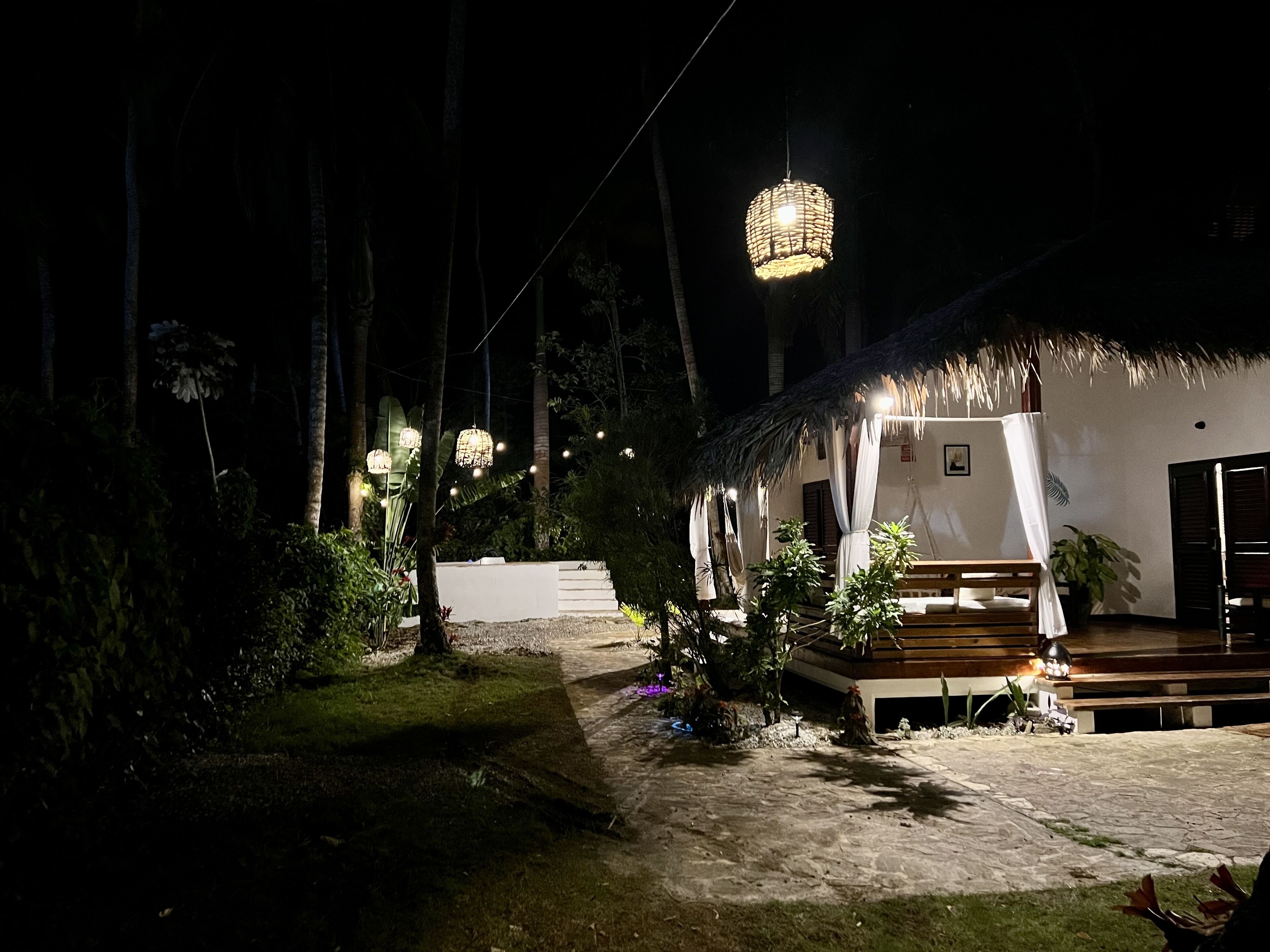 A thatched-roof outdoor area with hanging lights, a wooden deck, and a white building with a covered seating area.