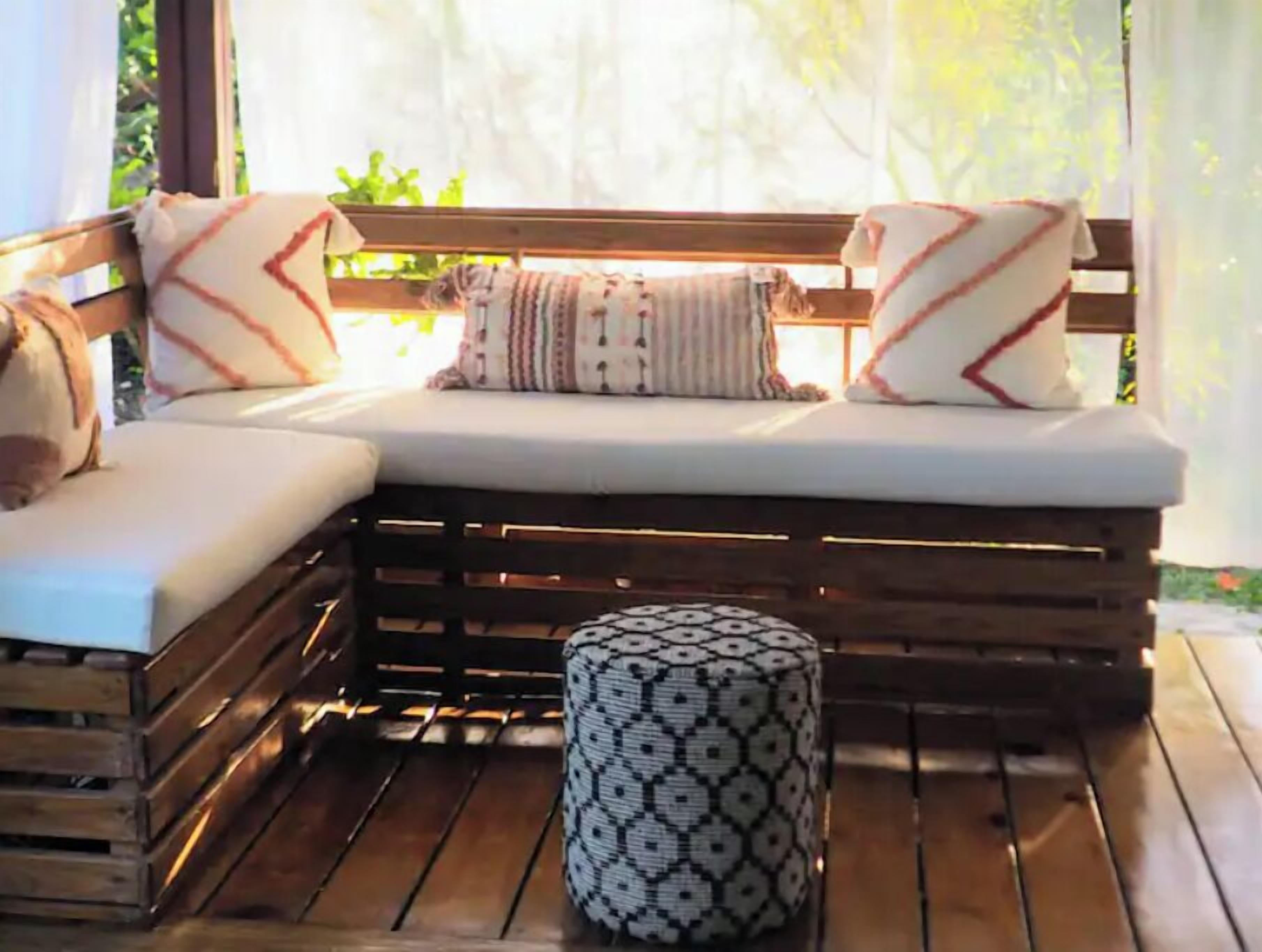 Une terrasse en bois avec un banc et un pouf, agrémentée d’un coussin et de plusieurs oreillers à motifs.