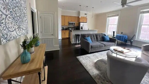 A spacious living area with a grey sofa, a wooden coffee table, and a blue rug. There is a kitchen in the background with wooden cabinets and a microwave. - Limited Offer: Cozy Condo by French Quarter (New Orleans)