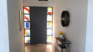 Un corridoio con una porta scura, un lampadario a parete, un tavolino con un vaso di fiori e uno sgabello in legno.