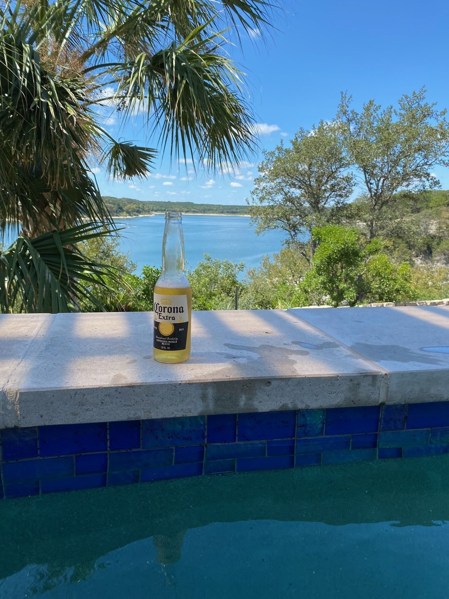 A bottle of Corona Extra beer on a poolside ledge with a view of a body of water and trees.