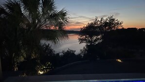 A swimming pool with a view of a sunset over water, surrounded by trees and a palm tree.