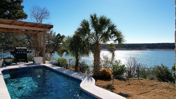 A swimming pool with a view of a lake and mountains, surrounded by palm trees and a barbecue area.