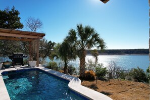 Una piscina con vistas a un lago y montañas, rodeada de palmeras y una zona de barbacoa.