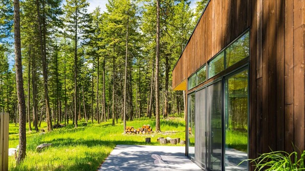 Ein modernes Holzgebäude mit großen Glasfenstern, umgeben von einem bewaldeten Gebiet mit einem deutlichen Weg zu einem Holzstapel.