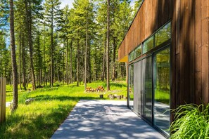 Un edificio moderno de madera con amplios ventanales, rodeado de un área boscosa con un sendero despejado que lleva a una pila de leña.