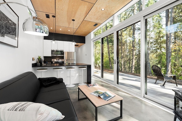 A modern kitchen with a central island, a dining area with a black sofa and a coffee table, and large windows offering a view of a forested area.