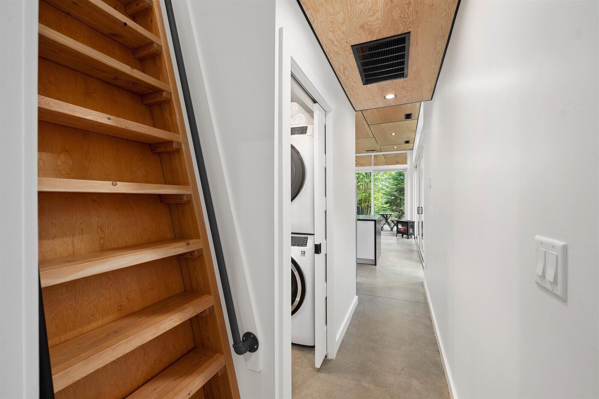 A modern interior with a wooden staircase, a white door leading to a laundry area, and a view of an outdoor garden.