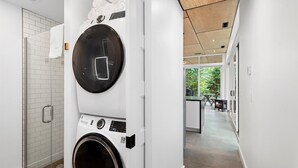 A modern laundry room with a large front-loading washer and dryer, a glass shower enclosure, and a view of a kitchen and outdoor area through a glass door. - The Clark Fork at Stoner Creek Cabins (Lakeside)