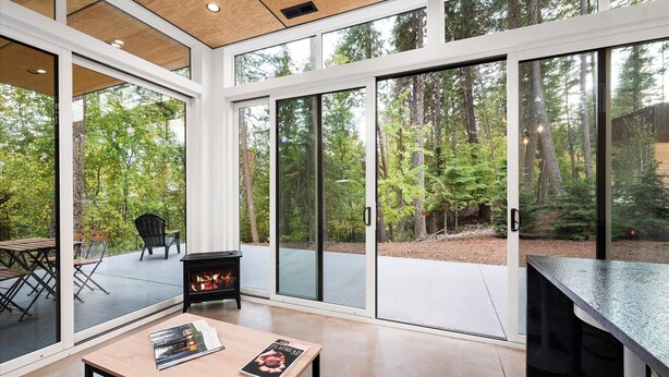 A modern interior with large sliding glass doors leading to a forested outdoor area, a fireplace, and a dining area with a table and chairs.