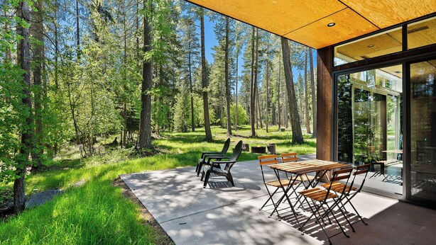 An outdoor patio with a concrete floor, wooden tables, and chairs, surrounded by a forested area. - The Bitterroot at Stoner Creek Cabins (Lakeside)