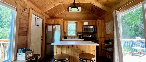 A wooden cabin interior with a kitchen area, two stools, and a large window.