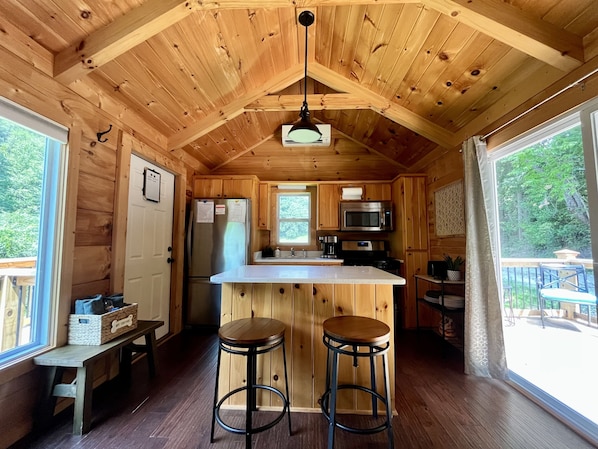 Interior de una cabaña de madera con una zona de cocina, dos taburetes y un ventanal grande.