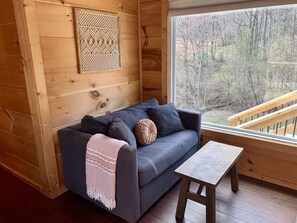 Interior acogedor de una cabaña con un sofá gris, una manta tejida y una mesa de madera. Un panel decorativo adorna la pared, y una ventana muestra vistas de un bosque.