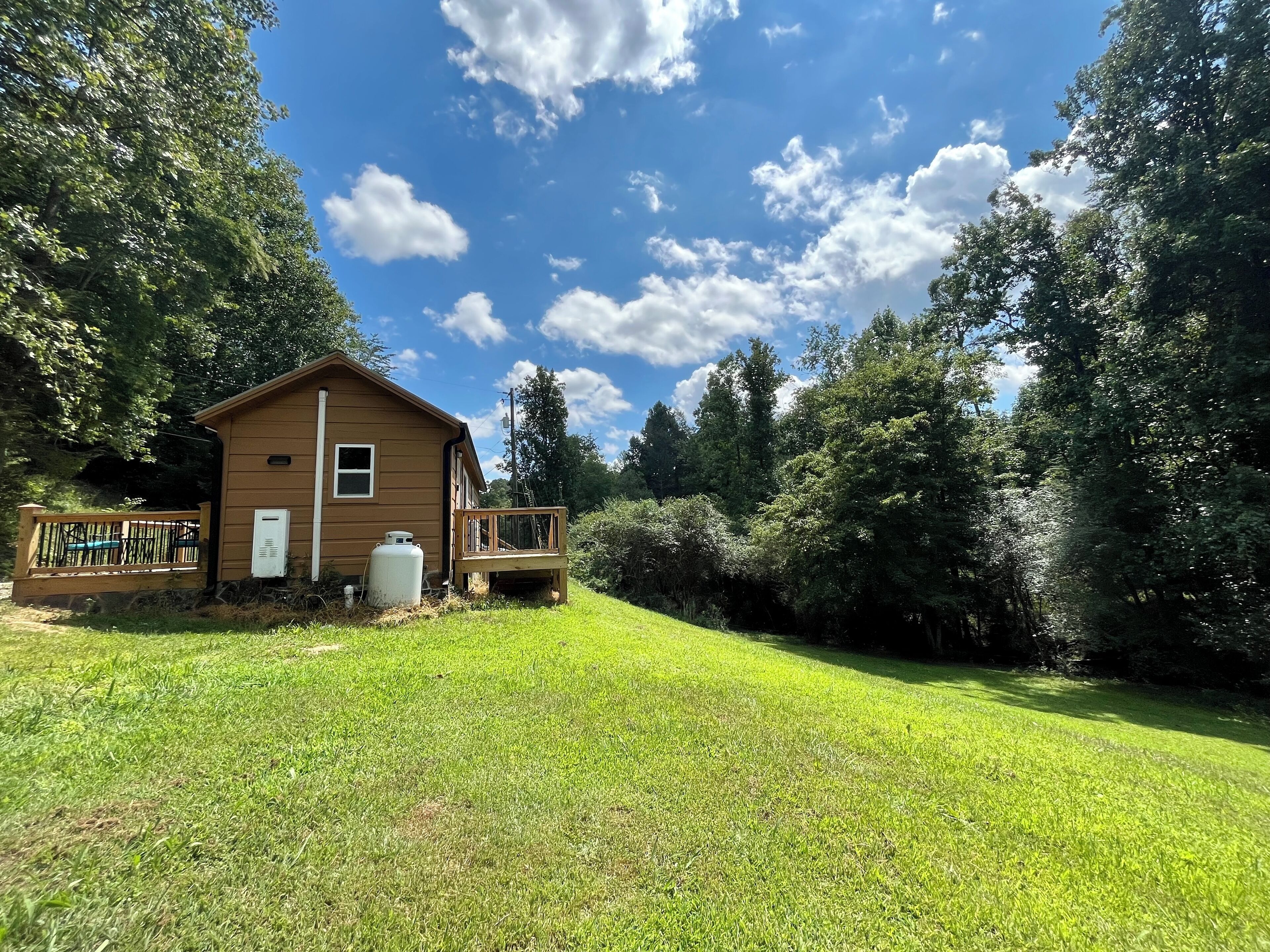 Une petite cabane en bois d’un seul étage, dotée d’une terrasse et entourée d’une végétation luxuriante et d’arbres.