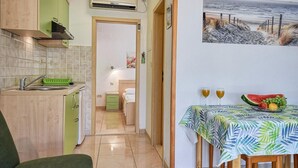A compact kitchen with a sink, a dining table with a fruit bowl, and a painting of a beach scene.