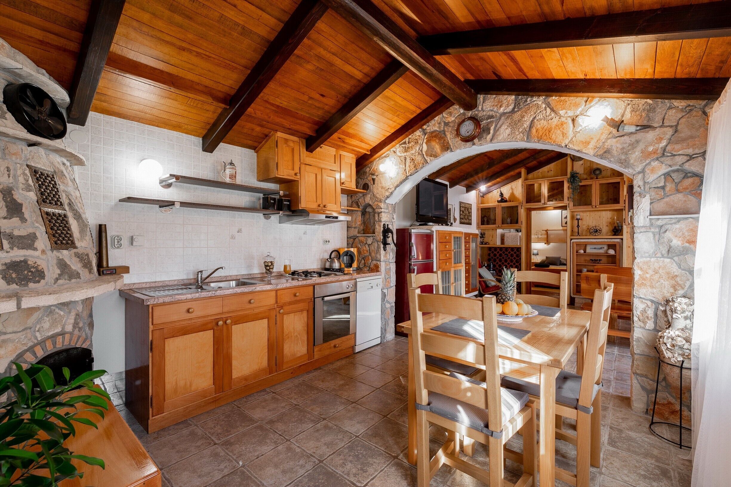 A rustic kitchen with wooden cabinets, a stone archway, and a dining area with wooden chairs.