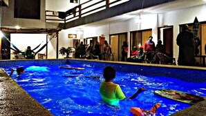 Zona de piscina con gente alrededor, un área de estar cubierta y un edificio con varias puertas y ventanas.
