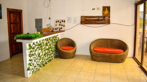 Un área de recepción con una puerta de madera, un mostrador con una planta, dos sillas de mimbre con cojines naranjas y una estantería con botellas.